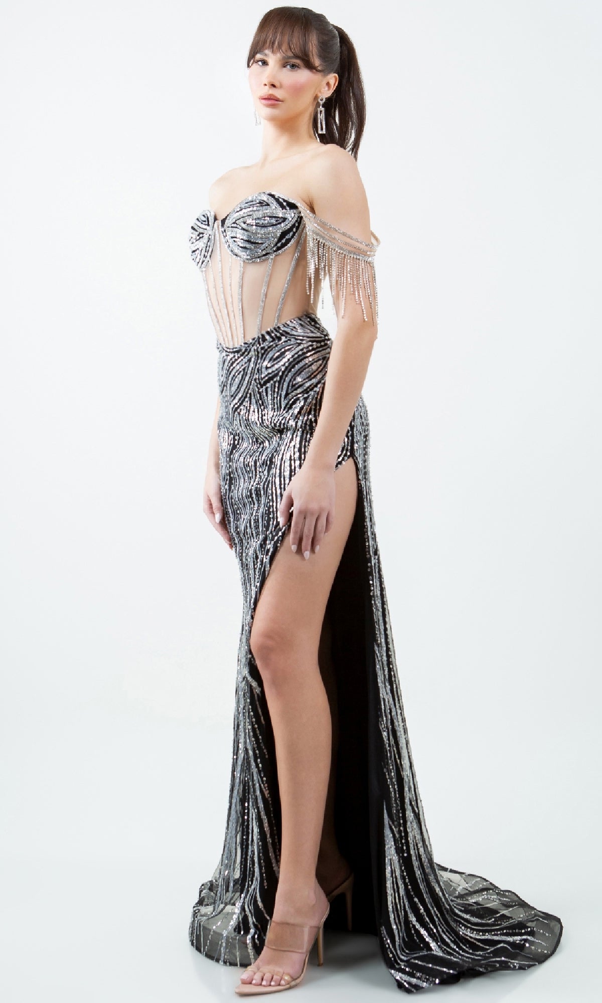 A woman models the Cinderella Couture 8147J long beaded prom dress in black and silver with a thigh-high slit and sheer corset bodice, paired with nude heels and drop earrings against a plain white background.